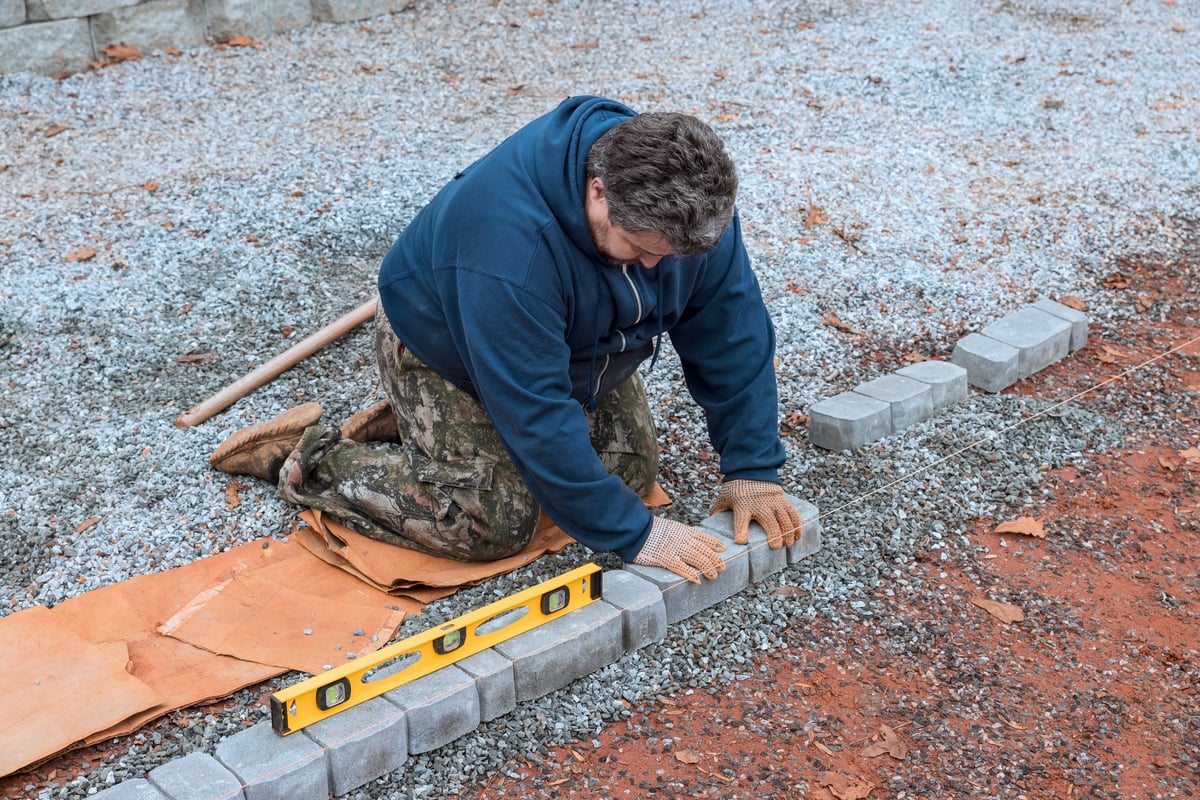 Worker carefully lays bricks for pathway, ensuring level placement on gravel surface during fall season.